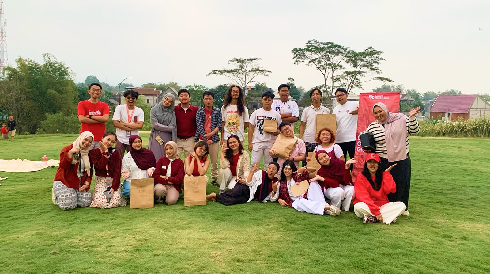 Indonesia Remote Club members gathered on an open lawn beside a red community banner, holding paper goodie bags